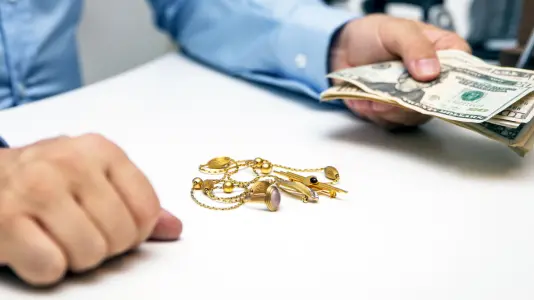 A closeup image of a man's hand holding a pile of USD and several gold jewelries on a table